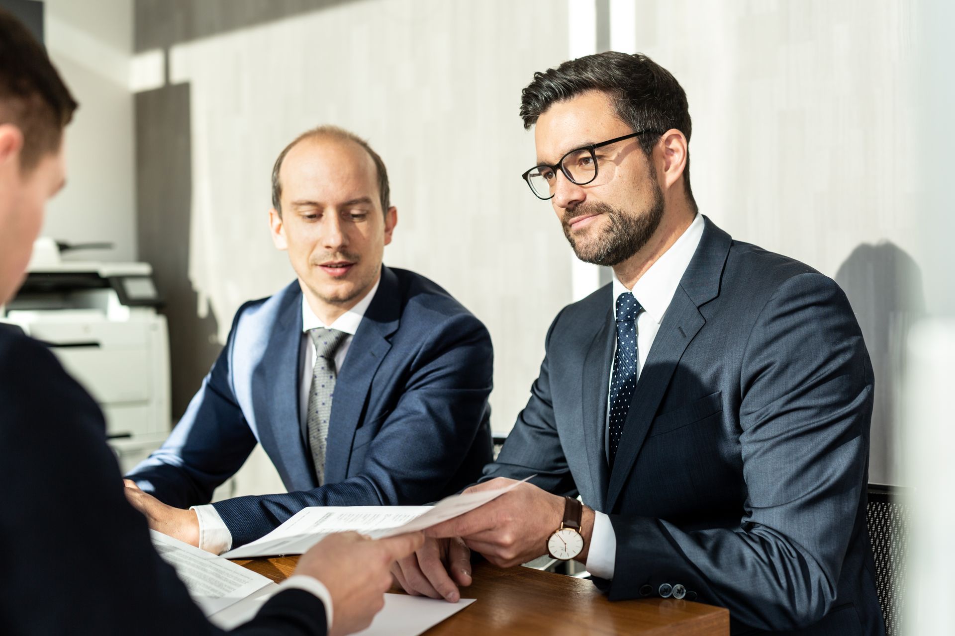 Three men in suits reviewing documents at a table in an office. 
