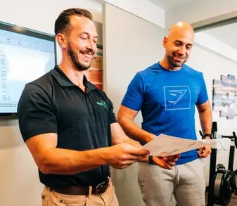 Two men smiling, looking at a paper in a gym setting. One wears a black polo, the other a blue tee.