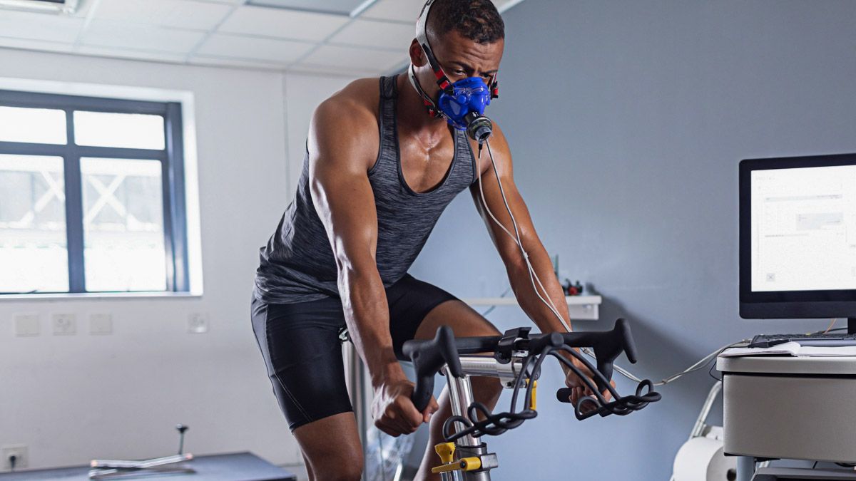 Man cycling indoors, wearing a mask, being monitored by equipment.