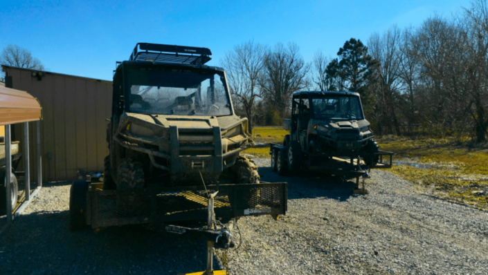 Two parked utility vehicles on gravel beside a tan building, with trees and blue sky in the background