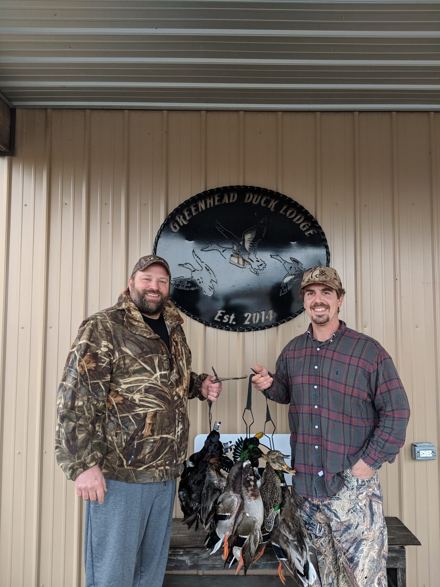 Two camo-clad hunters holding a pheasant and ducks beside a bird hunting lodge sign
