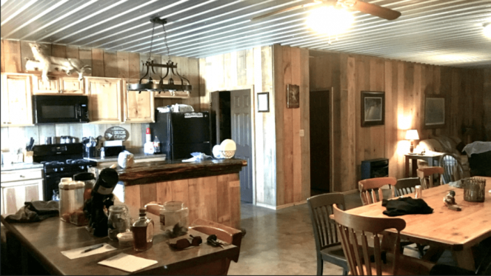 Rustic kitchen and dining area with wood-paneled walls, a long table, and a small black dog on the counter.