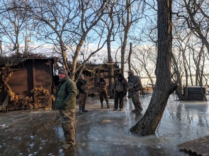 People standing on an icy outdoor path beside stacked firewood and leafless trees at dusk