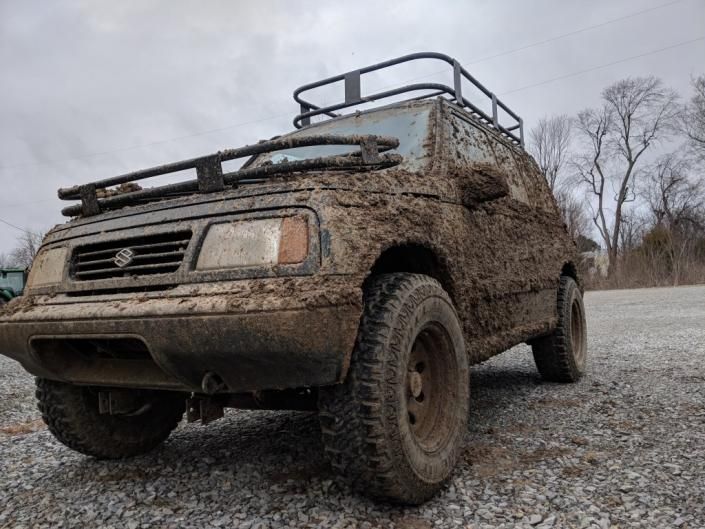 Mud-covered off-road SUV with roof rack parked on a gravel road under a cloudy sky