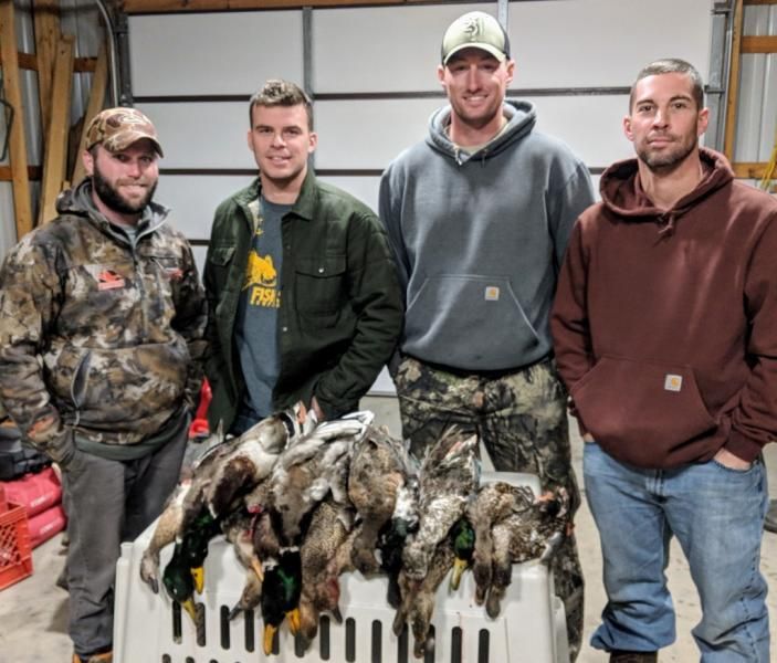 Four men stand behind a table piled with hunted ducks in a garage.