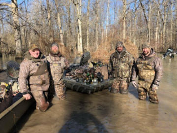 Camouflaged soldiers wade through muddy water with an armored vehicle in a wooded area