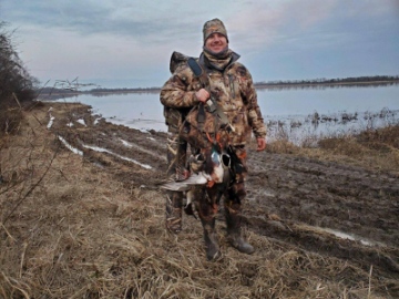 Person in camouflage holding several ducks on a muddy lakeshore in winter.