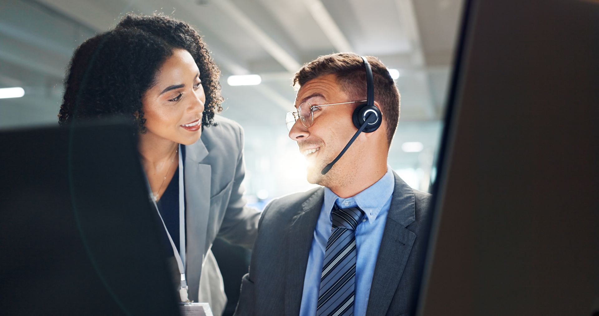 A man and a woman wearing headsets are looking at each other in front of a computer screen.