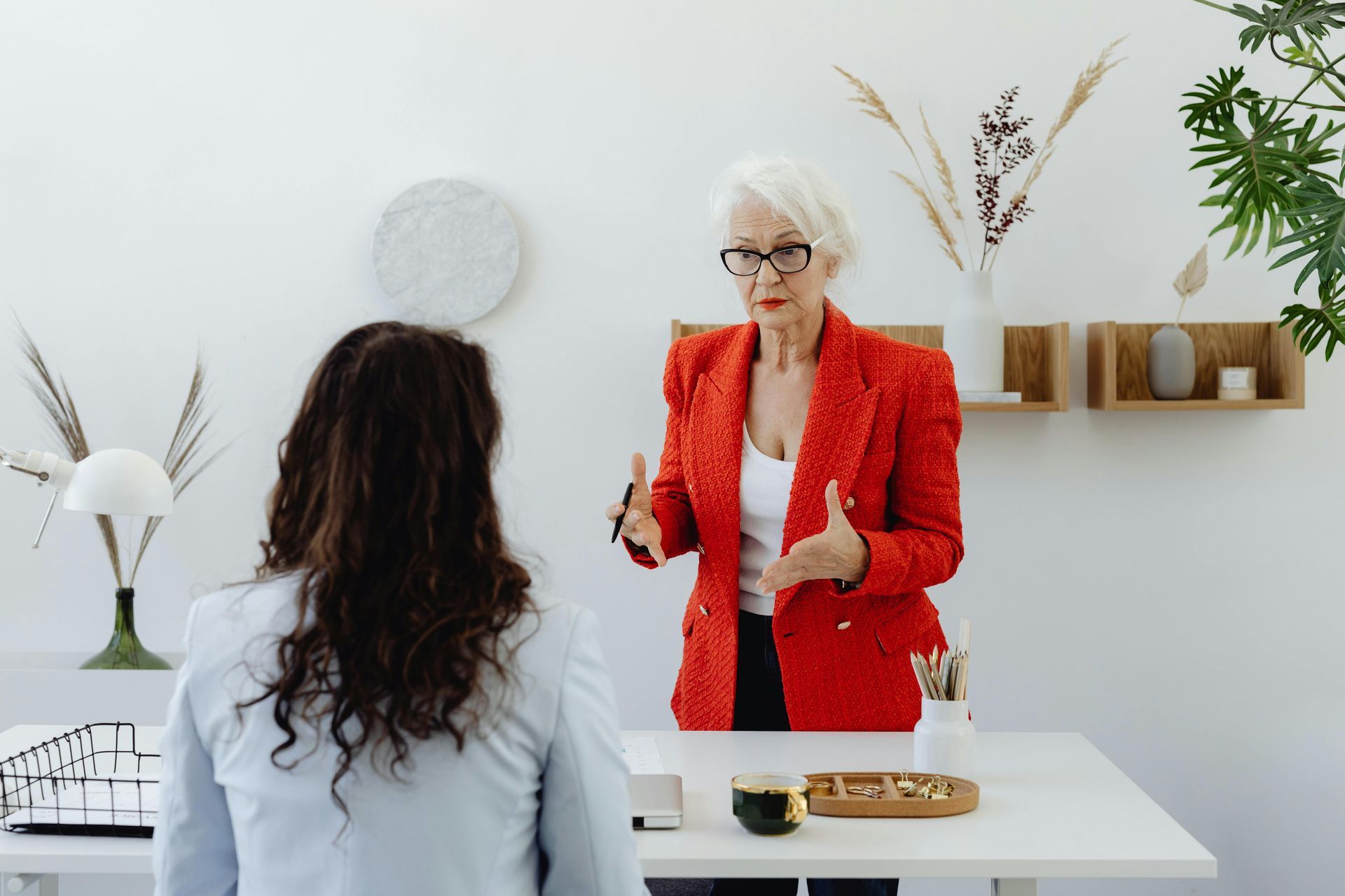 A woman in a red jacket is talking to a woman sitting at a table.