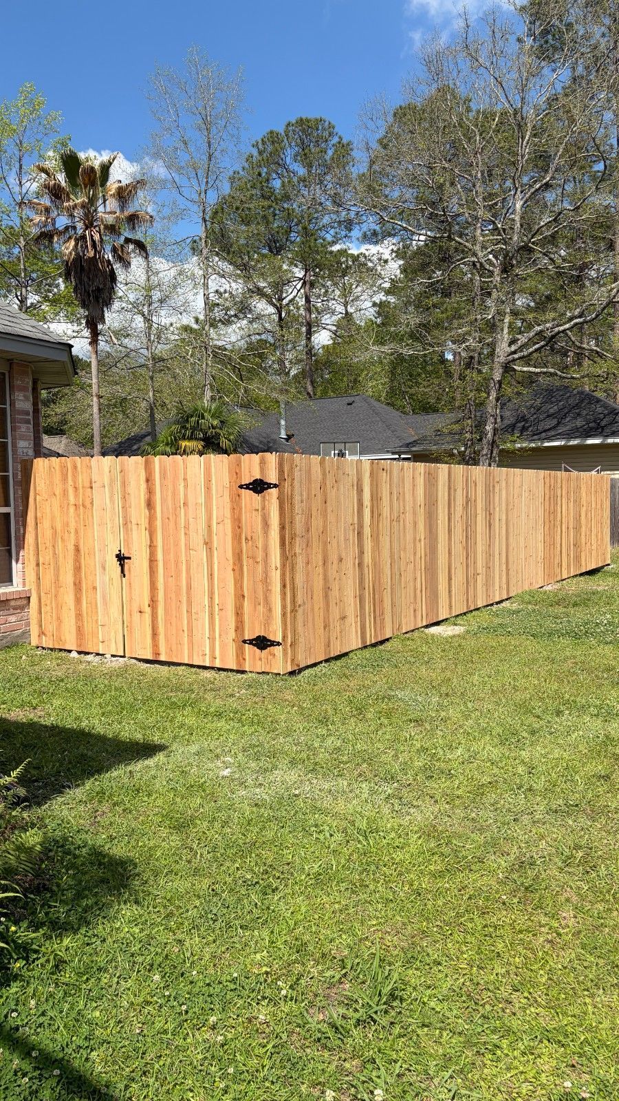 Wooden fenced-in yard enclosure on a grassy lawn, with trees and a house in the background.