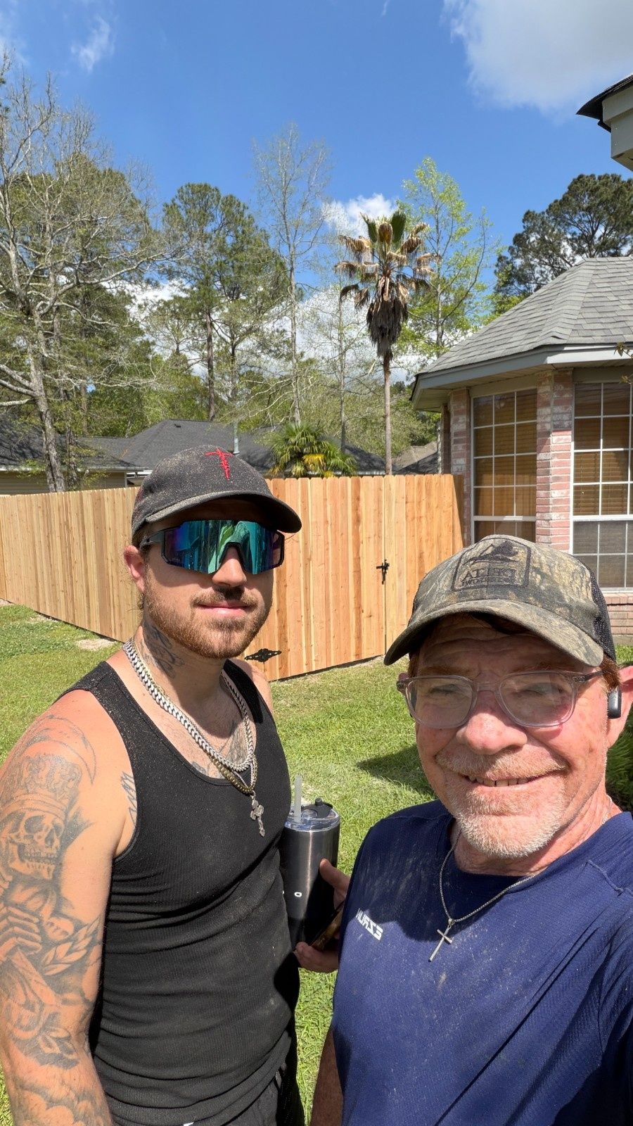 Two men smiling outdoors in a backyard, one taking a selfie in front of a wooden fence and house.