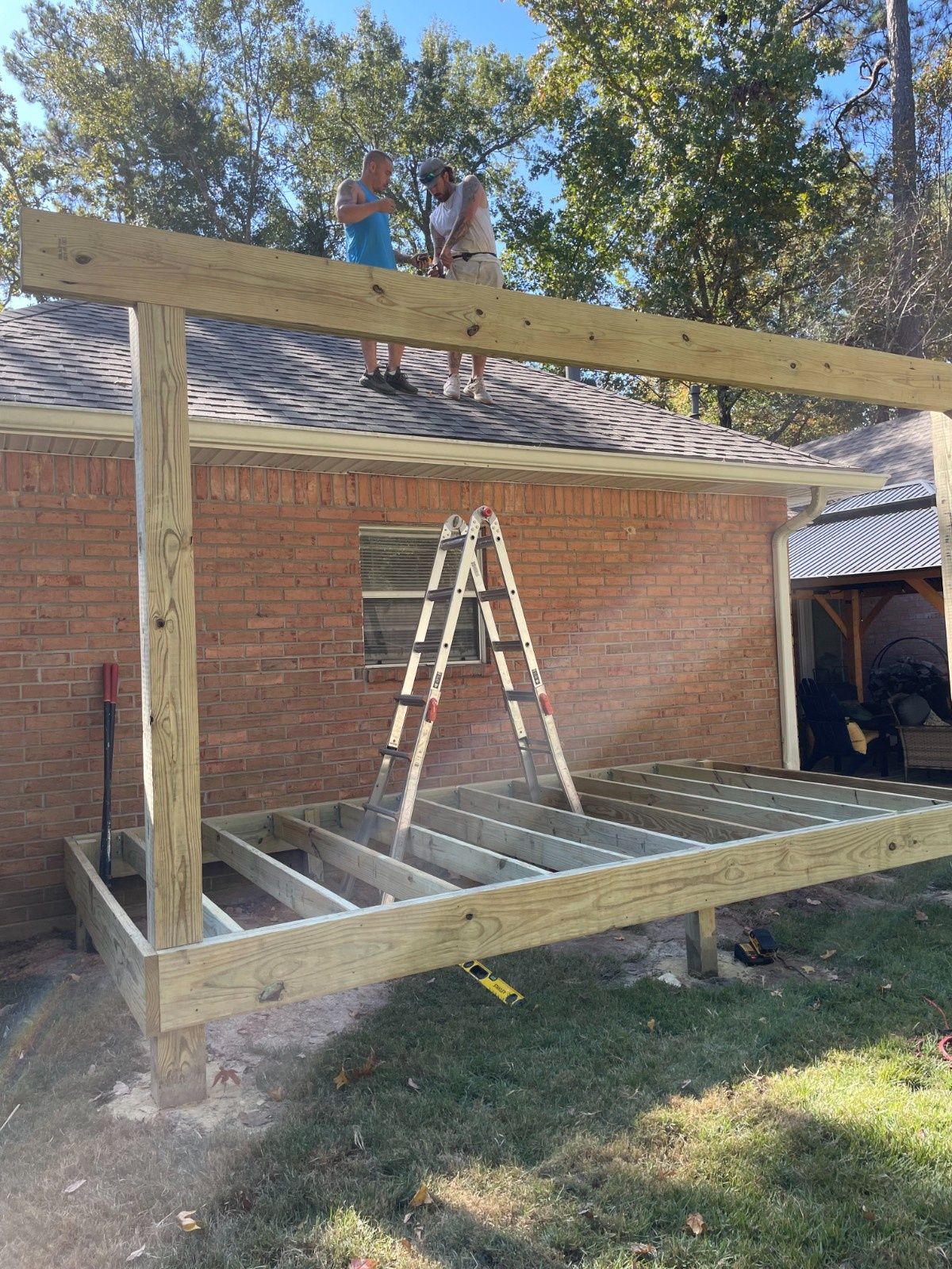 Person building a large wooden frame in a backyard beside a brick house, with a ladder inside.