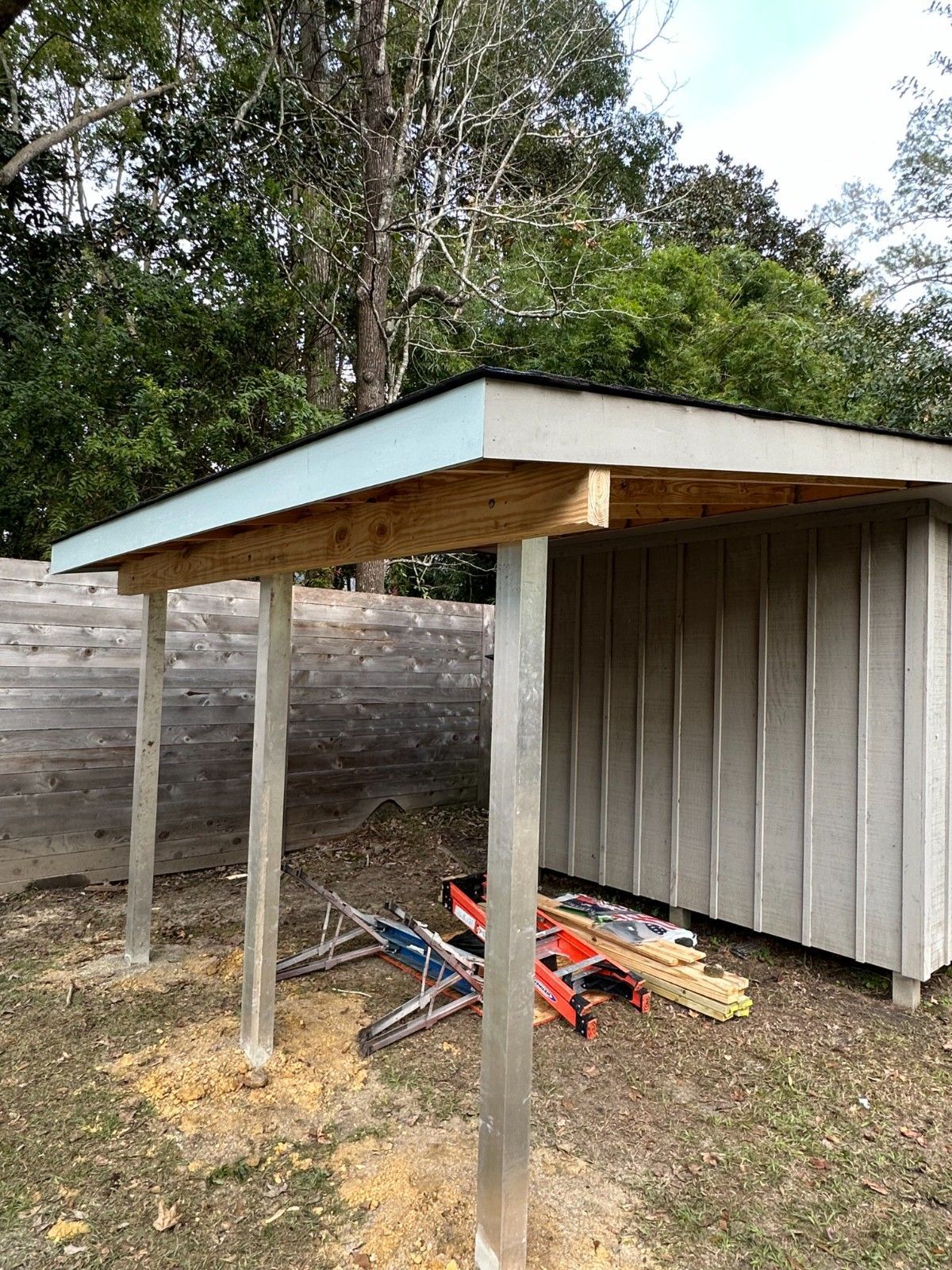 Simple outdoor shed with a slanted roof, supported by posts, beside a corrugated metal wall and tools on the ground.