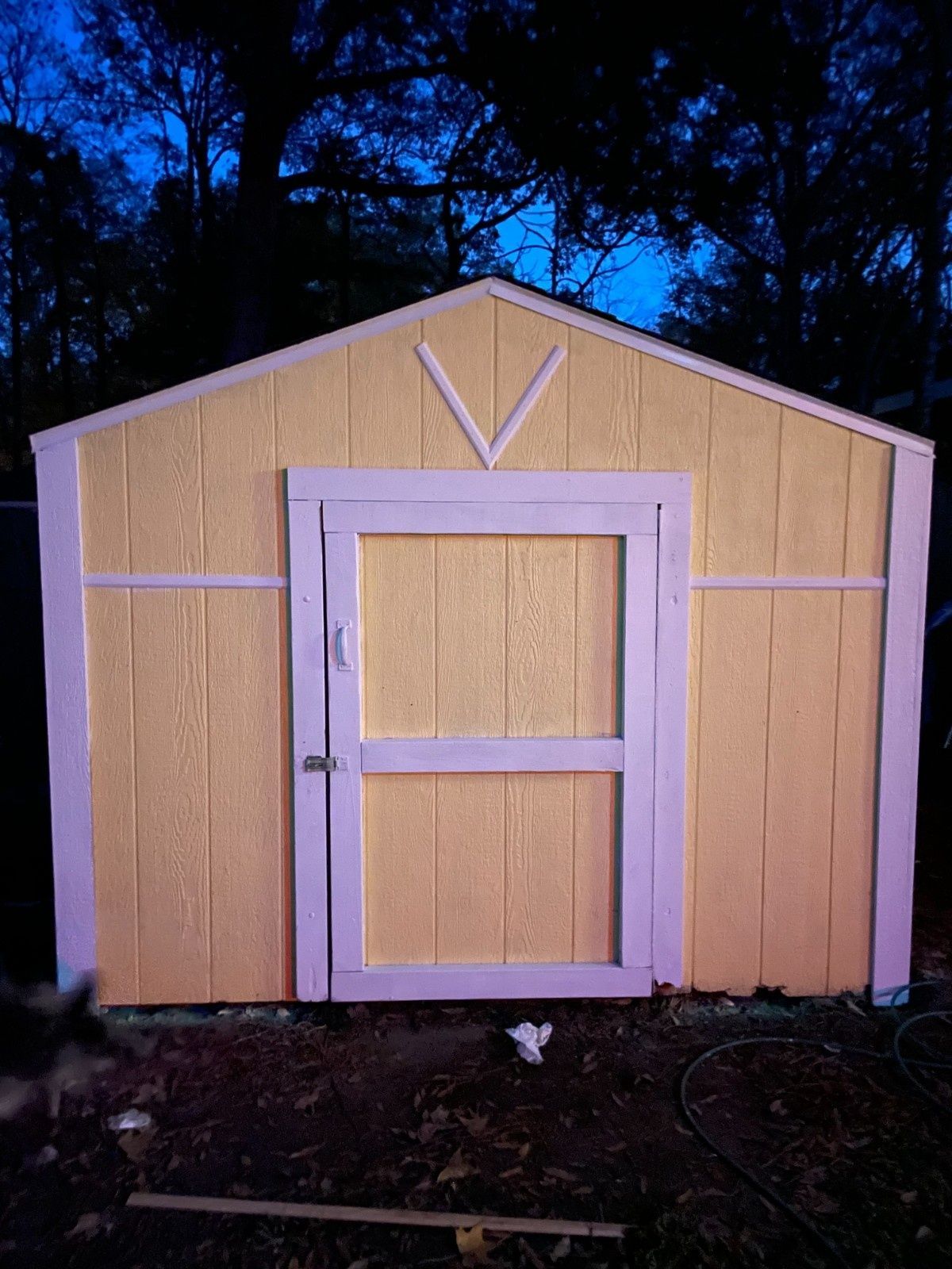 Small tan wooden shed with a white-framed door, lit against a dark outdoor background.