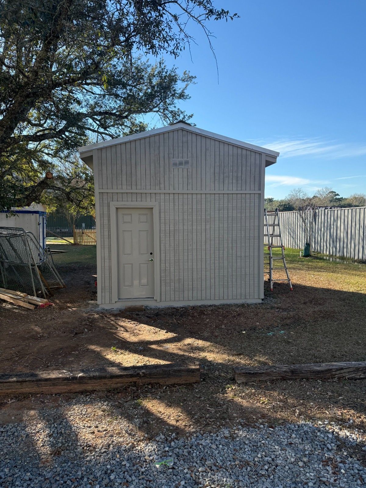 Small white shed with a door in a sunlit yard, trees and a fence in the background.