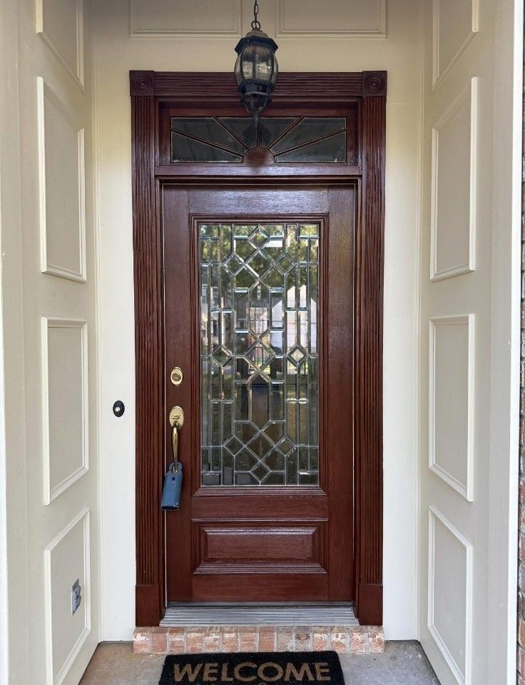 Front door with dark wood, glass panels, brass handle, and a welcome mat under a porch light.