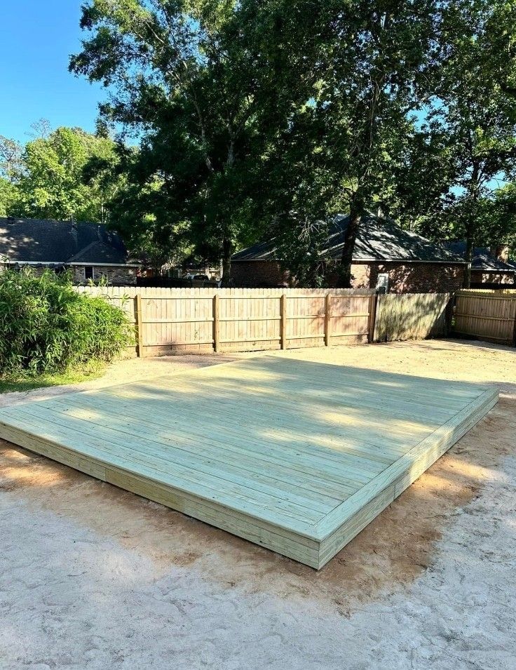Raised wooden platform in a fenced backyard under a large tree, with light-colored decking and dirt around it