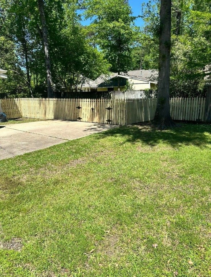 Sunny backyard with grass, a paved patio, and a wooden fence surrounded by trees.