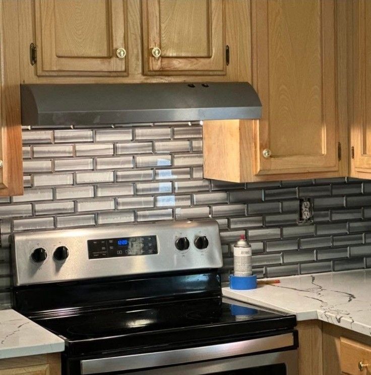 Kitchen stove with stainless steel range hood, wood cabinets, and gray tiled backsplash