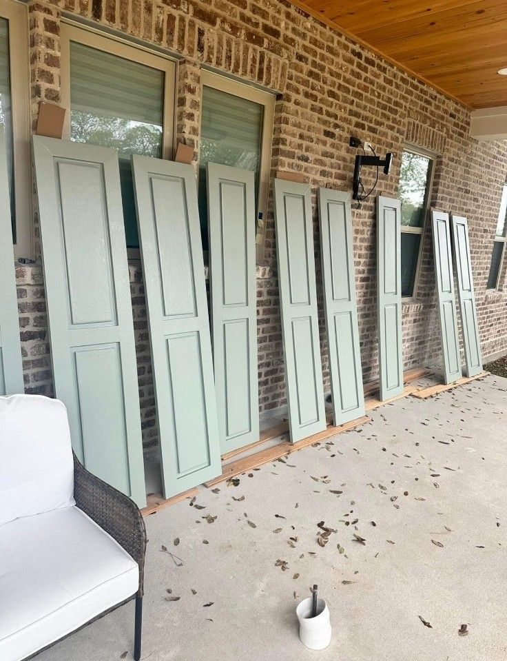 Pale green shutters and panels leaning against a brick porch wall under a wood ceiling.