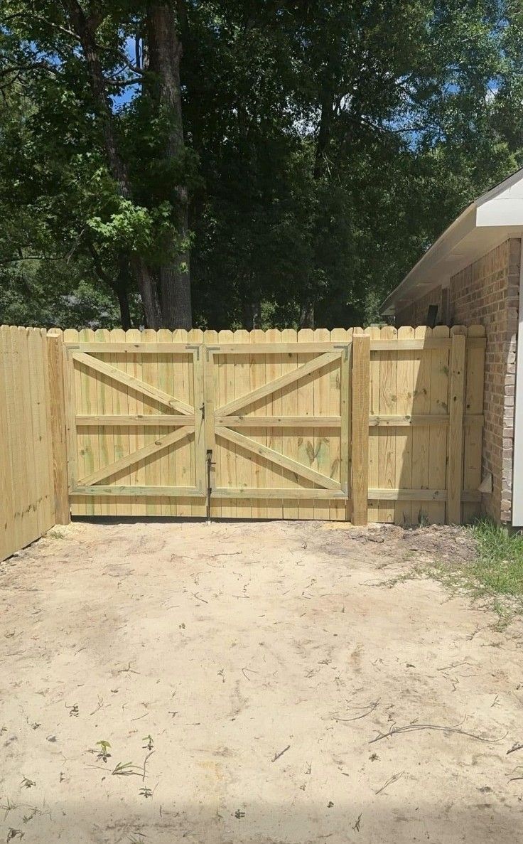 Wooden double gate in a fenced driveway, with trees and a house on the right.