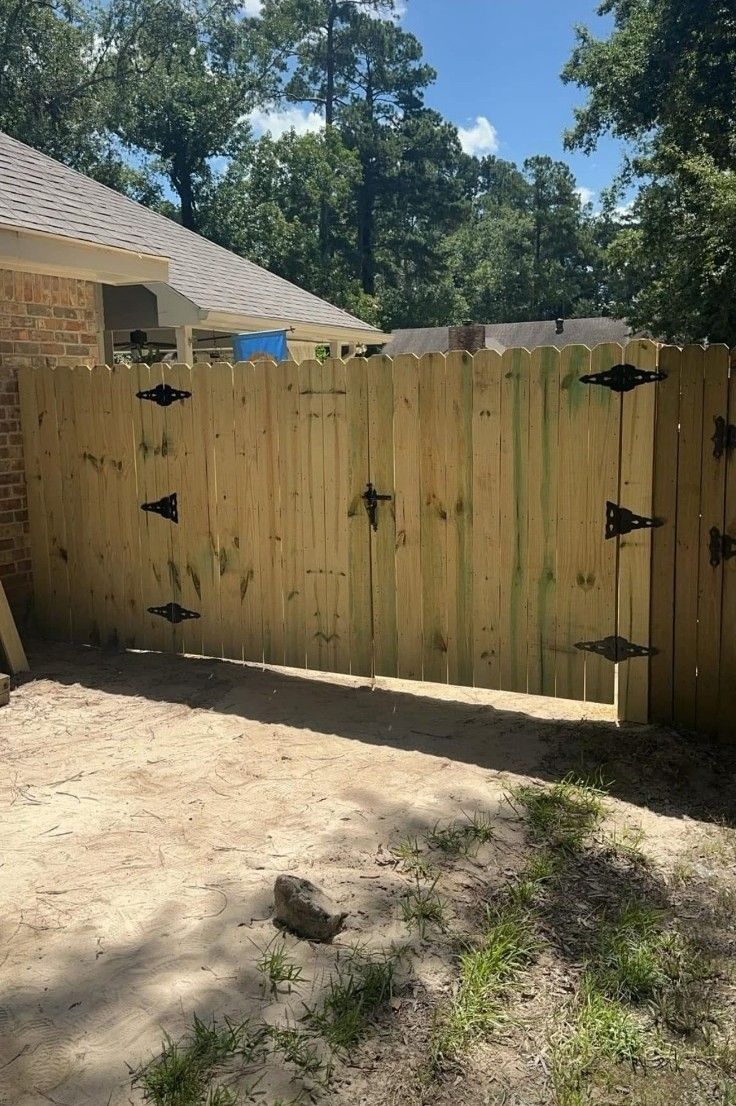 Wooden privacy gate with black hinges beside a house, opening to a sunny backyard with trees.