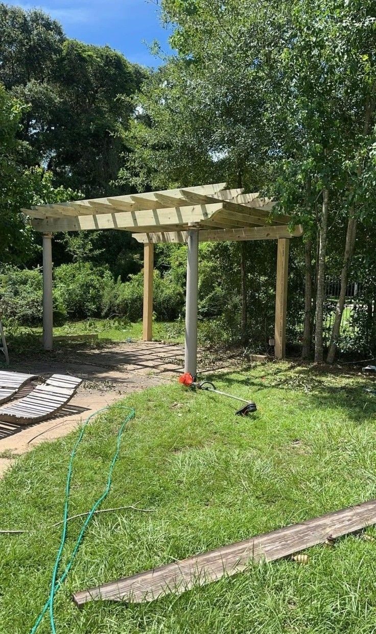 Wooden pergola in a sunny backyard with trees, grass, and a garden hose in the foreground