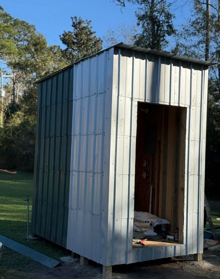 Small white metal shed with an open doorway, standing outdoors among trees and grass.