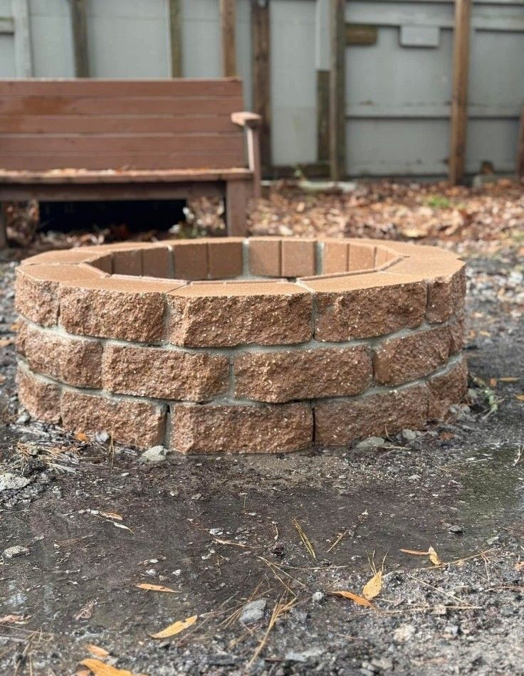 Round brick fire pit in a backyard, with a wooden bench and leaf-covered ground behind it