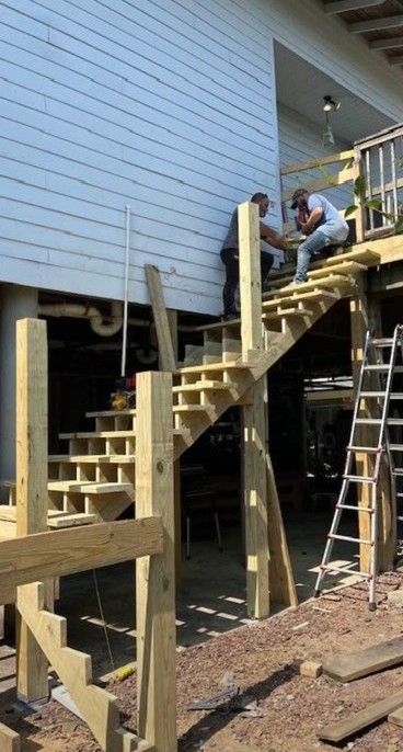 Workers building wooden stairs beneath a raised house, with one on the landing and a ladder nearby