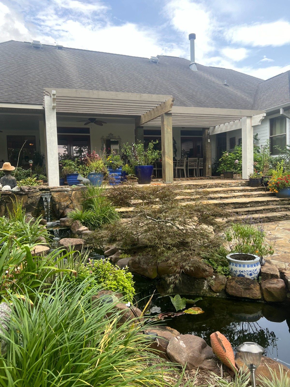 Garden patio with stone steps, pond, potted plants, and a white house with a wavy roof.