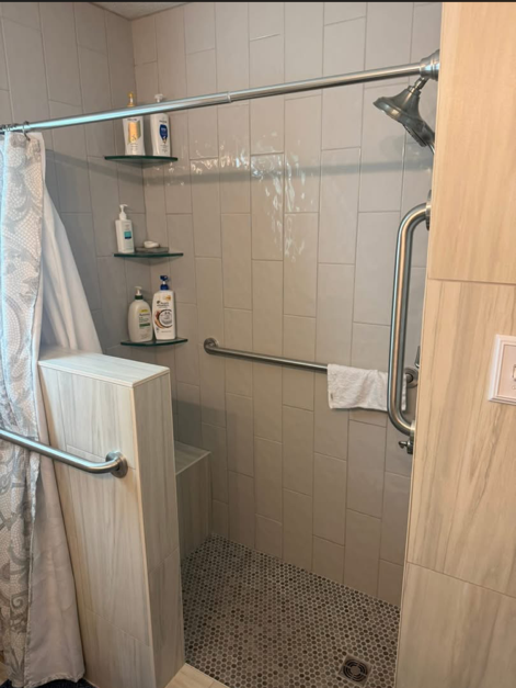 Small tiled shower stall with glass shelf, grab bars, and bath products.
