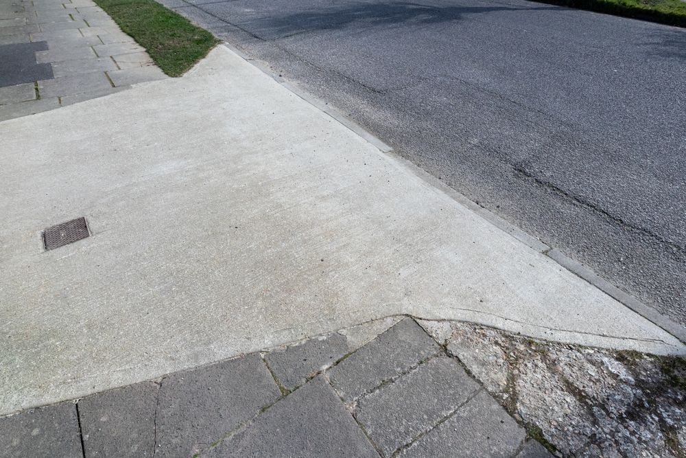 Cracked sidewalk corner beside an asphalt road with a curb drain and patchy grass.