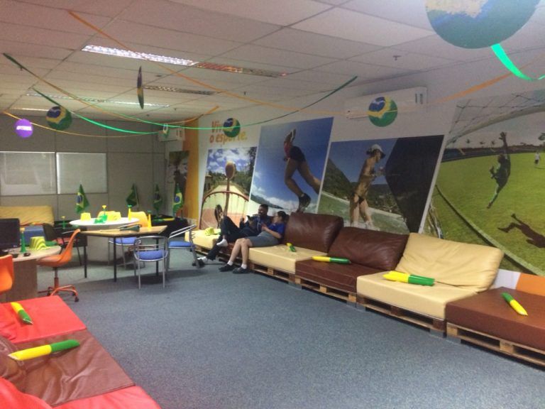 Uma sala decorada com as cores da bandeira brasileira, com pessoas descansando em sofás de paletes e mesas, algumas segurando pistolas de água.