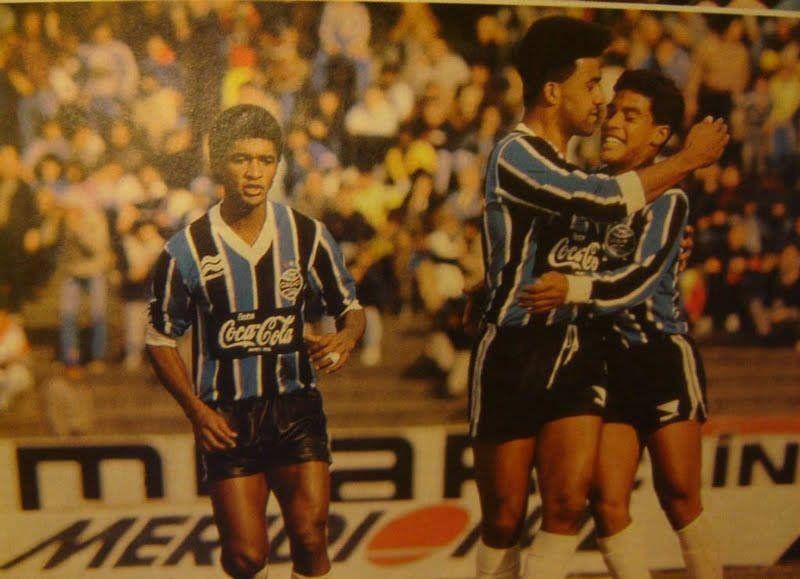 Jogadores de futebol com camisas listradas de azul e preto comemoram um gol em campo, enquanto outro jogador observa. A torcida ao fundo.