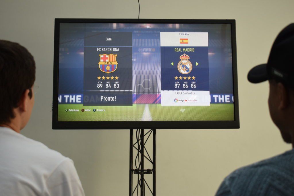 Duas pessoas jogando um videogame FIFA em uma tela grande. A configuração do jogo mostra uma partida entre Barcelona e Real Madrid.