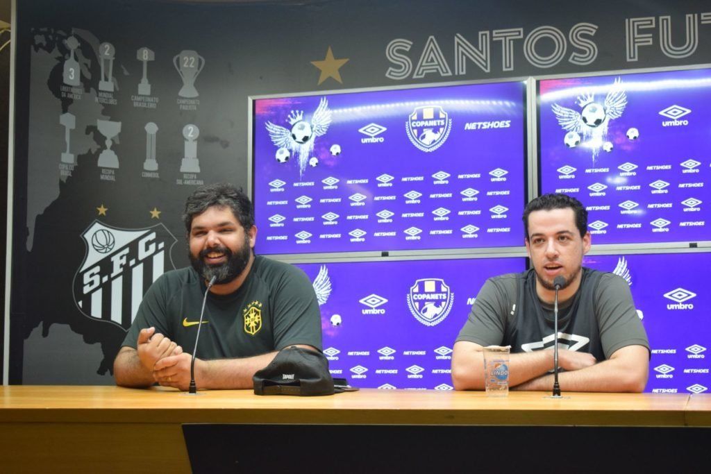 Dois homens estão sentados à mesa, possivelmente em uma coletiva de imprensa. Atrás deles, há um cenário com o logotipo do Santos FC. O homem à esquerda sorri.