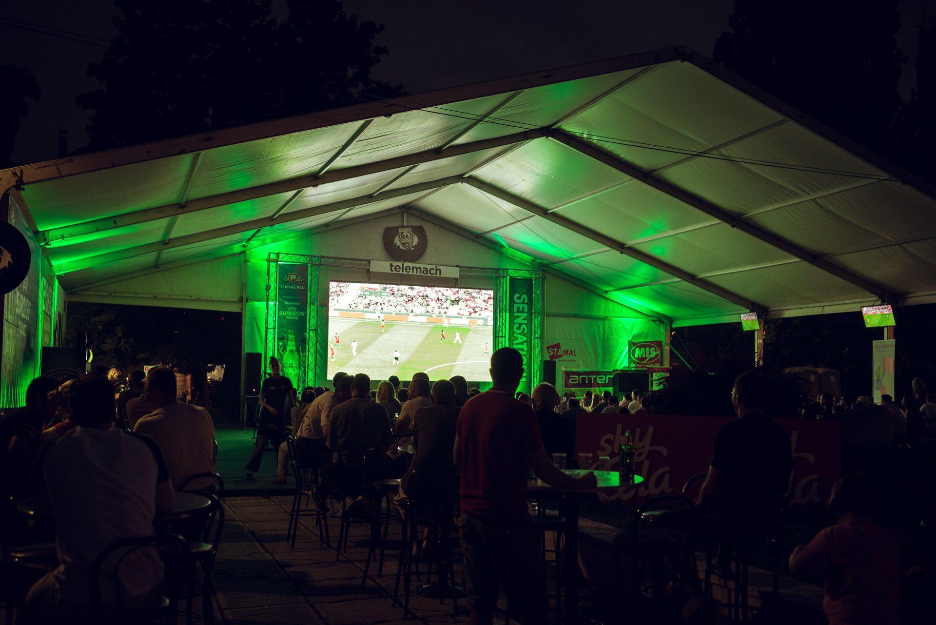 Pessoas assistindo a um jogo esportivo em uma tela grande dentro de uma tenda iluminada com luzes verdes à noite.