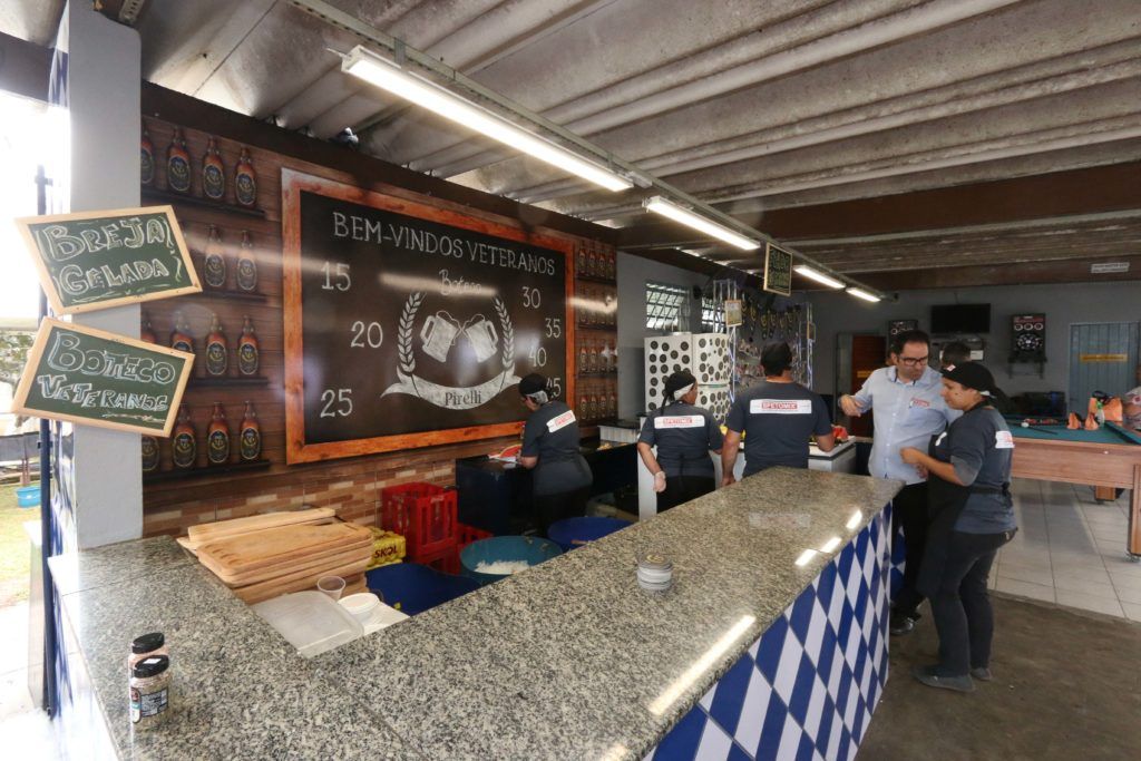 Um bar com funcionários, clientes e um design xadrez. O bar é ao ar livre, com uma parede de tijolos e uma placa de lousa com o nome do bar.