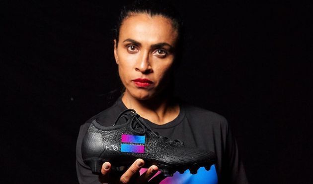 Mulher de cabelos escuros segurando um sapato preto com detalhes coloridos. Ela está em um ambiente escuro, olhando para a câmera.