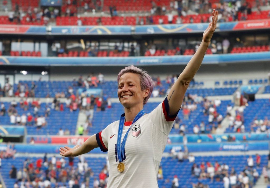 Megan Rapinoe, com uma medalha de ouro, levanta os braços em comemoração em um campo de futebol. Ela veste uma camisa da Seleção Feminina dos EUA com o estádio ao fundo.