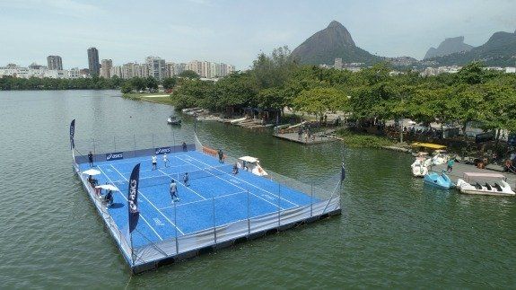 Uma quadra azul flutuante em um lago sedia uma partida de tênis de praia no Rio de Janeiro, com o Pão de Açúcar ao fundo.