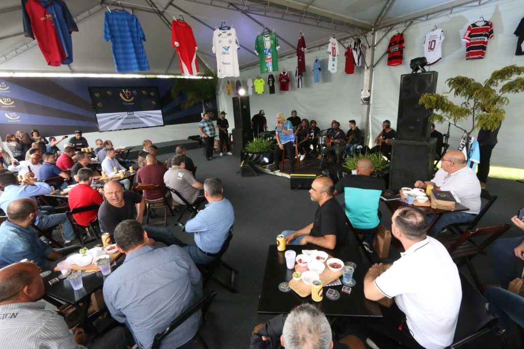 Pessoas reunidas dentro de uma tenda, socializando em mesas. Camisas esportivas penduradas no teto; uma banda ao fundo.