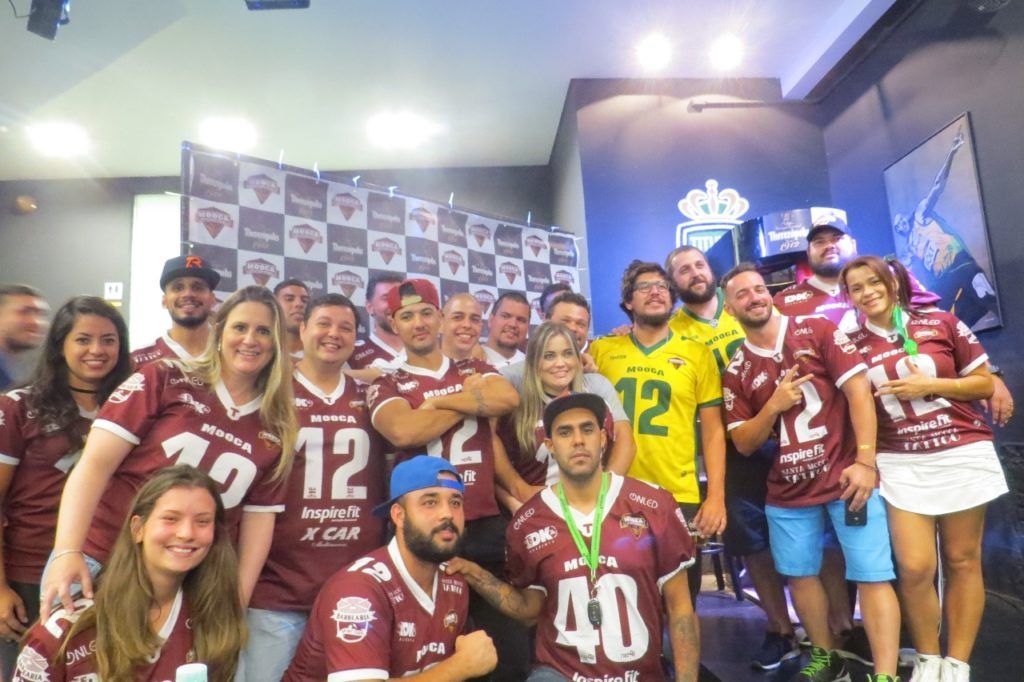Grupo de pessoas com camisas bordô posando para uma foto, algumas abraçadas. Um homem veste uma camisa amarela. Eles estão em um ambiente interno com um logotipo ao fundo.