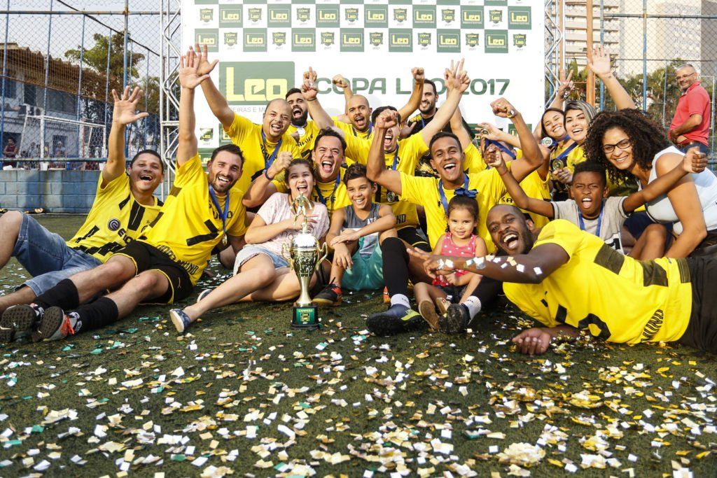 Um time de futebol, vestindo camisas amarelas e pretas, comemora a vitória, cercado por confete e com os braços erguidos. Eles estão em um campo com uma faixa ao fundo.