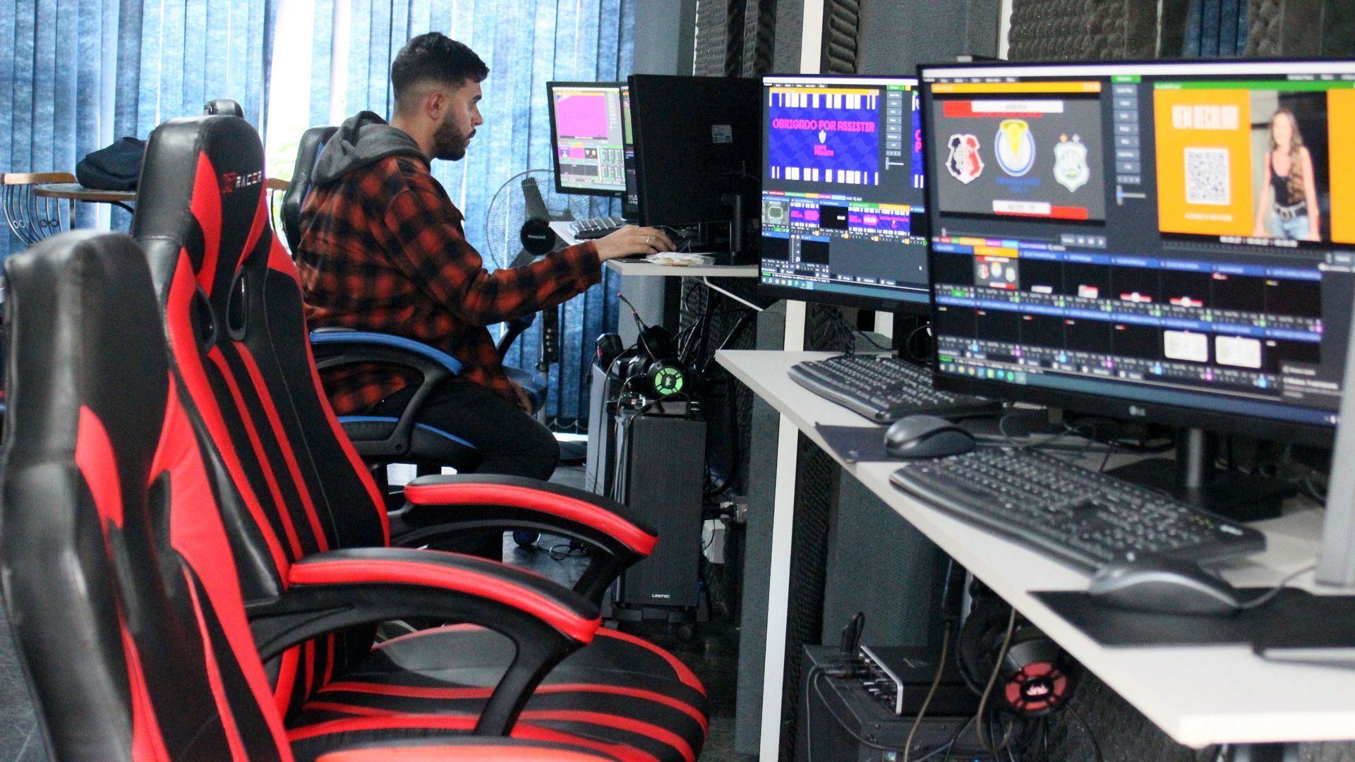 Um homem de camisa xadrez trabalha em um computador em um estúdio. Dois monitores são visíveis, juntamente com uma cadeira gamer vermelha e preta.