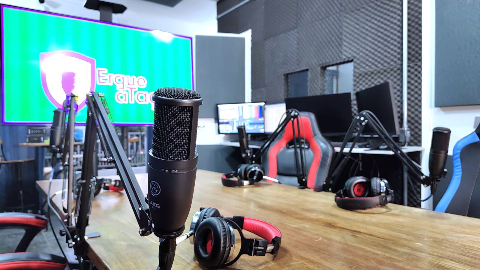 Estúdio de gravação com microfones, fones de ouvido, monitores e uma tela de fundo. Uma mesa de madeira e uma cadeira gamer também aparecem na cena.