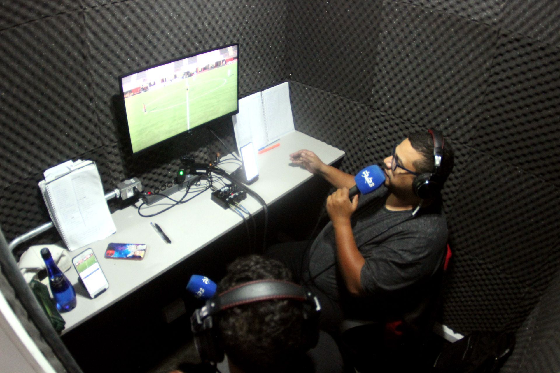 Duas pessoas em uma cabine à prova de som, transmitindo. Um homem de óculos segura um microfone e olha para um monitor exibindo uma partida de futebol.