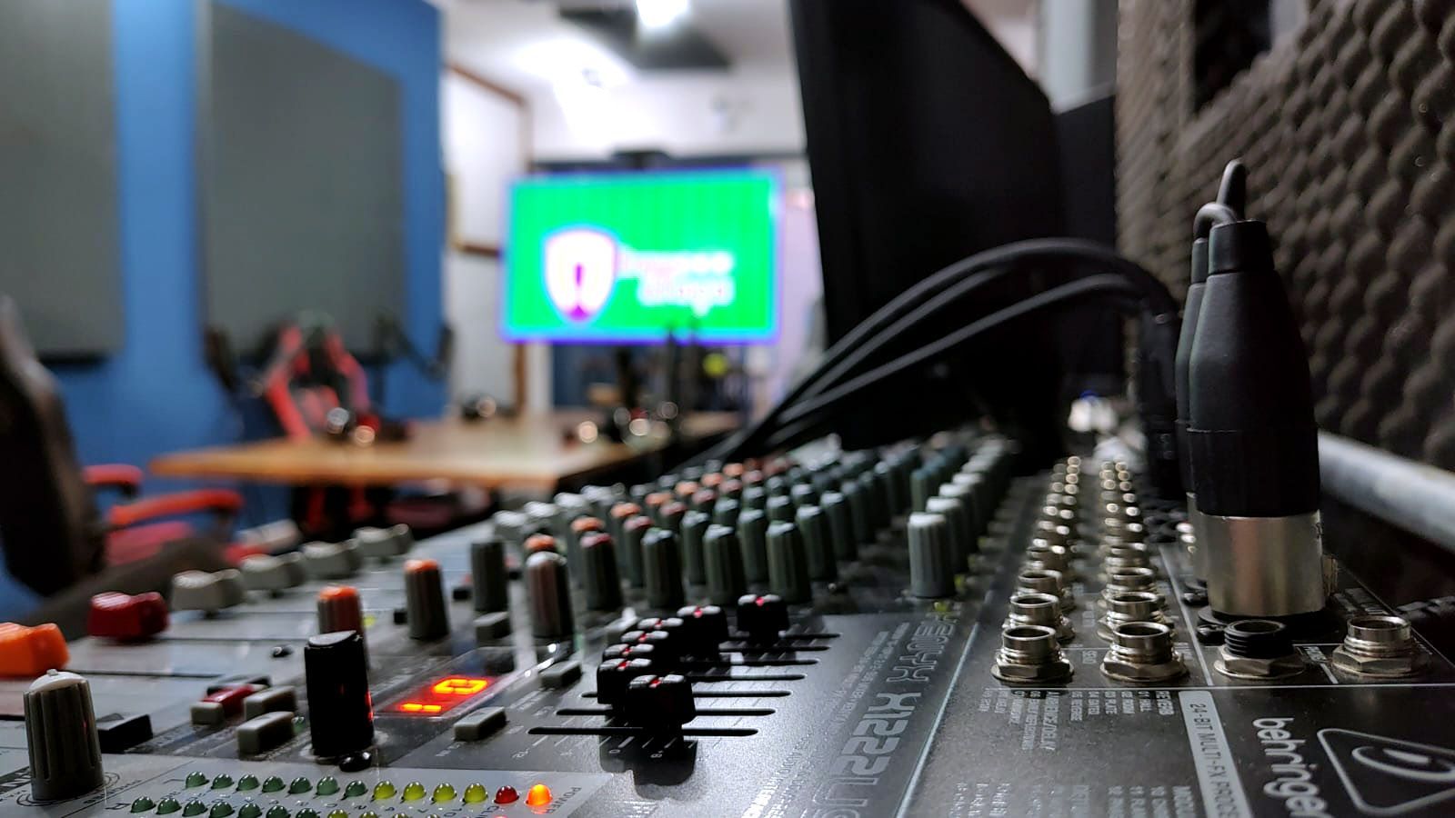 Close-up de uma mesa de mixagem de som com botões e cabos, uma tela exibindo um logotipo ao fundo e um cenário de estúdio de gravação.