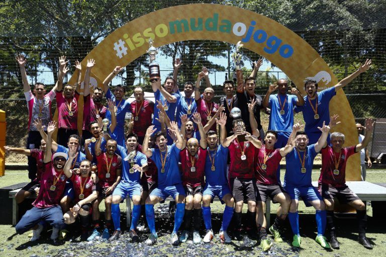 Time de futebol com uniforme azul e marrom comemorando uma vitória ao ar livre sob um arco com o texto